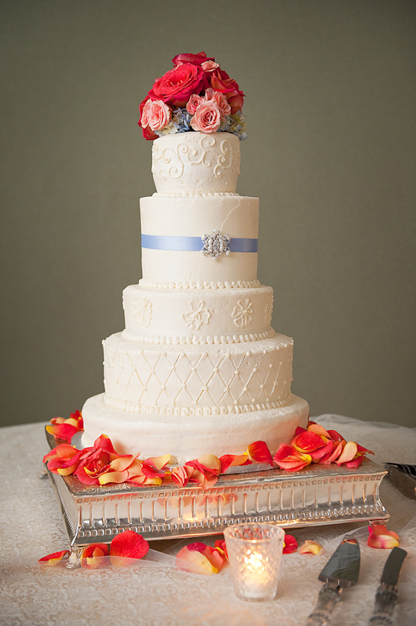 image of a beautiful white five tier round wedding cake decorated with a light blue ribbon and silver brooch and a coral, pink, and blue floral topper - cake is on a silver stand decorated with coral, pink, and yellow rose petals - photo by Houston based wedding photographer Adam Nyholt 